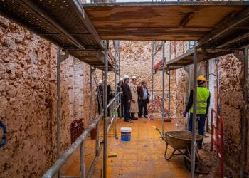 Un gruppo di persone che indossano caschi e indumenti di sicurezza si trovano sul pavimento piastrellato giallo di un cantiere del tribunale di Albufeira con impalcature e muri di pietra non finiti. In primo piano sono visibili una carriola e materiali da costruzione.