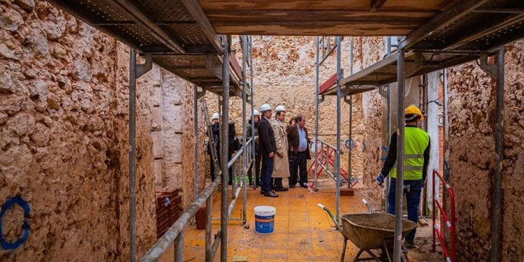 Un gruppo di persone che indossano caschi e indumenti di sicurezza si trovano sul pavimento piastrellato giallo di un cantiere del tribunale di Albufeira con impalcature e muri di pietra non finiti. In primo piano sono visibili una carriola e materiali da costruzione.