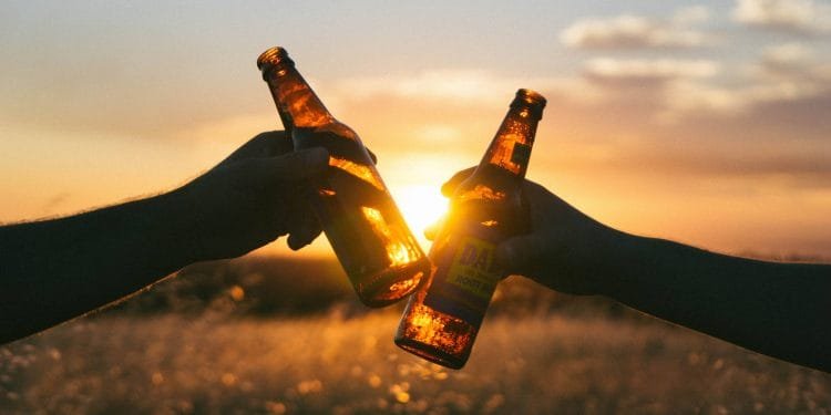Due mani tengono bottiglie di birra che tintinnano insieme sullo sfondo del sole al tramonto. La calda luce del tramonto illumina le bottiglie, creando un'atmosfera rilassata e celebrativa. La scena si svolge in un campo aperto con erba alta.