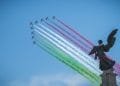 Una formazione di aerei emette scie di fumo rosso, bianco e verde, che rappresentano i colori della bandiera italiana, contro un cielo azzurro e limpido. In primo piano è visibile la statua scura di un angelo. Sono le Frecce Tricolori mentre suona Inno di Mameli.