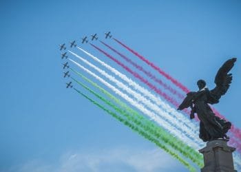 Una formazione di aerei emette scie di fumo rosso, bianco e verde, che rappresentano i colori della bandiera italiana, contro un cielo azzurro e limpido. In primo piano è visibile la statua scura di un angelo. Sono le Frecce Tricolori mentre suona Inno di Mameli.
