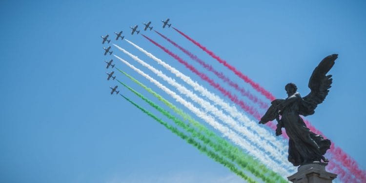 Una formazione di aerei emette scie di fumo rosso, bianco e verde, che rappresentano i colori della bandiera italiana, contro un cielo azzurro e limpido. In primo piano è visibile la statua scura di un angelo. Sono le Frecce Tricolori mentre suona Inno di Mameli.