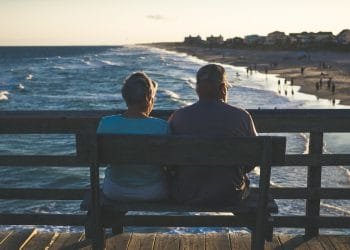 Una coppia di anziani, post-coronavirus, siede vicina su una panchina su un molo, di fronte all’oceano. Godono di una vista tranquilla sulla spiaggia, sulle onde ondeggianti e sulle persone che camminano lungo la costa al tramonto. Il cielo è leggermente nuvoloso.