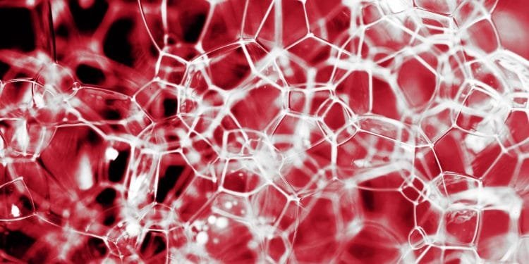 Un primo piano di bolle di sapone intricate e interconnesse con uno sfondo rosso cremisi, che creano un motivo simile a una ragnatela. Le bolle sono traslucide e di dimensioni variabili, conferendo all'immagine un aspetto delicato e astratto.