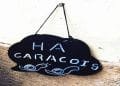 Un cartello nero con scritte bianche è appeso a un muro. Si legge "HÁ CARACÓIS" e presenta due lumache disegnate a mano nella parte inferiore. Il cartello è fissato al muro con un solo chiodo.