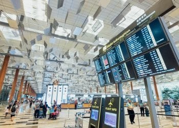 Un terminal aeroportuale spazioso con soffitti alti e design architettonico moderno. I passeggeri camminano con i carrelli portabagagli. In alto è appeso un grande tabellone con le informazioni sui voli che mostra gli orari di arrivo e partenza. Si possono vedere anche chioschi touchscreen e aree salotto.