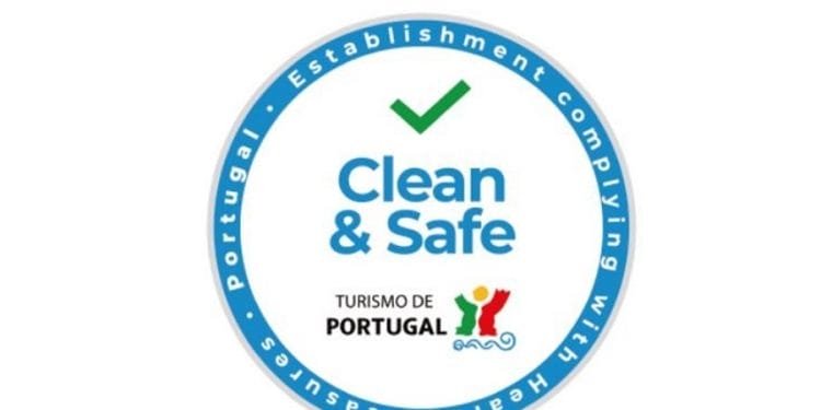Un badge circolare con la scritta "Clean and Safe" con un segno di spunta verde sopra. Il testo che circonda il badge dice "Stabilimento conforme alle misure sanitarie - Turismo de Portugal". Vengono inoltre visualizzate l'icona di una figura colorata e le onde. Questo è il logo della Piattaforma digitale Clean & Safe.
