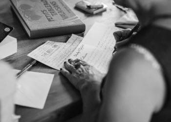 Immagine in bianco e nero di una persona seduta a un tavolo di legno, che scrive una lettera a mano. Ci sono più fogli, una penna, una busta, un libro intitolato "PETITS BEAUX VALISES" e un piccolo oggetto sul tavolo. La scena cattura un momento intimo che ricorda la prima uscita in pizzeria di qualcuno.