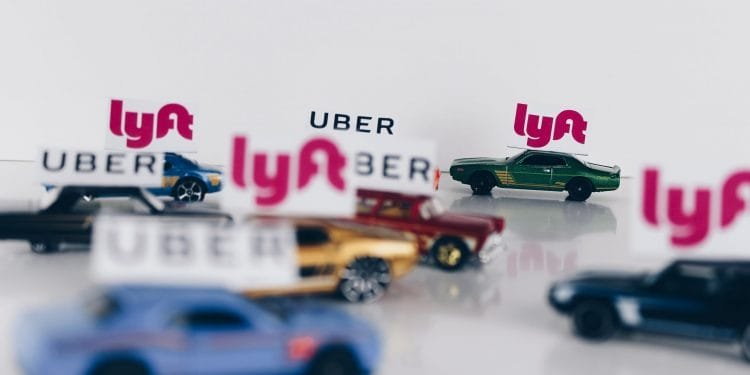 Un gruppo di piccole macchinine con segnaletica Uber e Lyft sui tetti disposte su una superficie bianca. Le auto sono di vari colori e i segnali appaiono ben in vista nell'immagine. I loghi Lyft sono in magenta, mentre i loghi Uber sono in nero. I conducenti Uber sono TVDE.