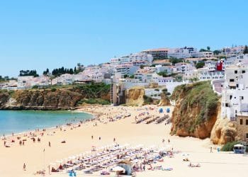 Spiaggia soleggiata con sabbia dorata, fiancheggiata da sedie a sdraio e ombrelloni bianchi. Le persone prendono il sole e nuotano nell'acqua cristallina. Scogliere con case ed edifici in cima si affacciano sulla spiaggia. Il cielo è limpido e azzurro, il che rende facile capire perché Albufeira è leader tra le destinazioni balneari.