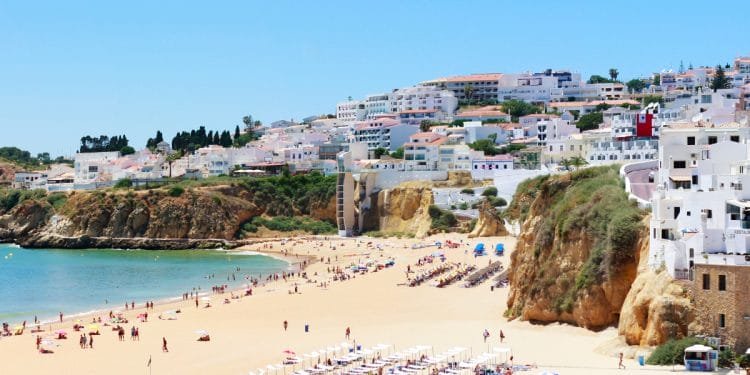 Spiaggia soleggiata con sabbia dorata, fiancheggiata da sedie a sdraio e ombrelloni bianchi. Le persone prendono il sole e nuotano nell'acqua cristallina. Scogliere con case ed edifici in cima si affacciano sulla spiaggia. Il cielo è limpido e azzurro, il che rende facile capire perché Albufeira è leader tra le destinazioni balneari.
