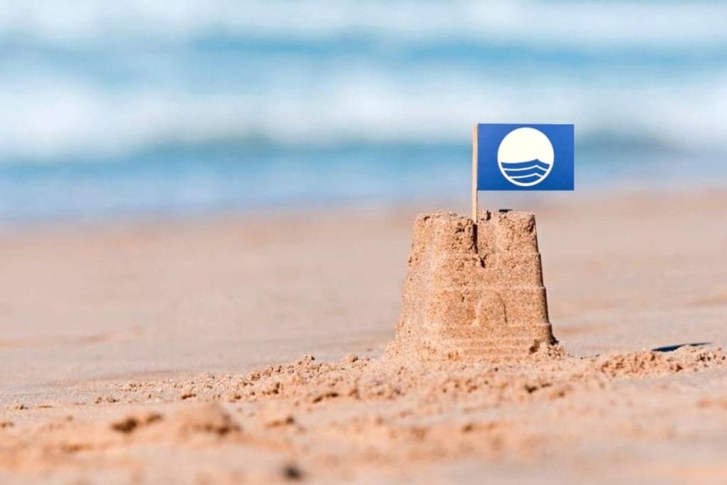 Un piccolo castello di sabbia su una spiaggia con una piccola bandiera blu con un'onda bianca e un disegno circolare. Le onde dell'oceano si confondono dolcemente sullo sfondo, mentre il castello di sabbia è messo a fuoco in primo piano.