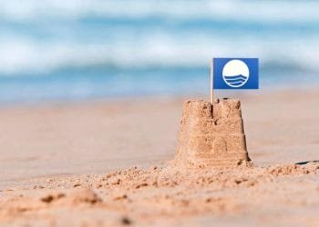 Un piccolo castello di sabbia su una spiaggia con una piccola bandiera blu con un'onda bianca e un disegno circolare. Le onde dell'oceano si confondono dolcemente sullo sfondo, mentre il castello di sabbia è messo a fuoco in primo piano.