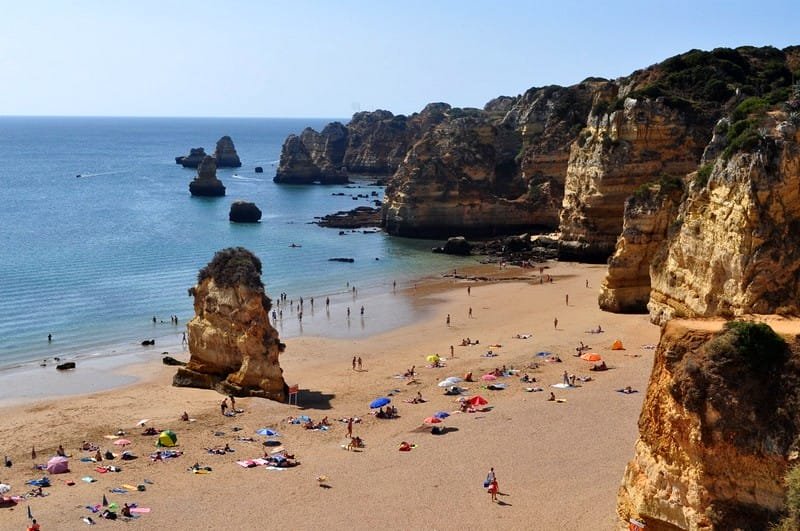 Una spiaggia panoramica in Algarve con sabbia dorata, acqua cristallina e scogliere rocciose. Numerose persone prendono il sole, nuotano e si rilassano sotto ombrelloni colorati. Le scogliere si protendono nell'oceano, formando piccole insenature e pittoresche formazioni rocciose nell'acqua.