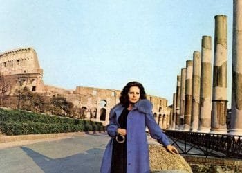 Una donna che è Amália Rodrigues che indossa un cappotto blu si trova di fronte a un sito storico con alte colonne di pietra e il Colosseo sullo sfondo. La scena è ambientata in una giornata limpida, con l'antica struttura e il verde visibili nell'area circostante.