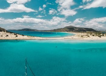 Veduta aerea di un sereno paesaggio costiero caratterizzato da vivaci acque turchesi, spiagge di sabbia bianca e lontane aspre colline sotto un cielo parzialmente nuvoloso. Le barche sono sparse sull'acqua vicino alla riva, esaltando la scena idilliaca.