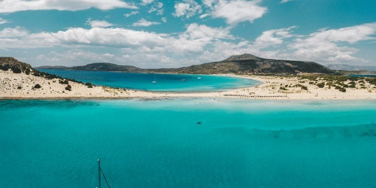 Veduta aerea di un sereno paesaggio costiero caratterizzato da vivaci acque turchesi, spiagge di sabbia bianca e lontane aspre colline sotto un cielo parzialmente nuvoloso. Le barche sono sparse sull'acqua vicino alla riva, esaltando la scena idilliaca.