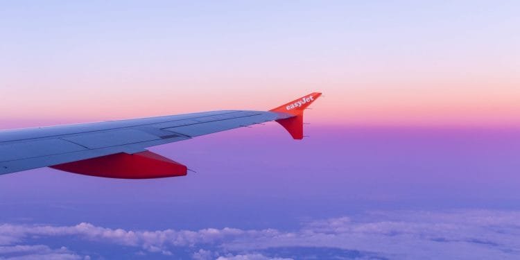 Vista dal finestrino di un aereo che mostra un vivace cielo al tramonto rosa e viola. L'ala e l'ala dell'aereo, marchiate con "easyJet" in testo bianco su sfondo arancione, si estendono nell'inquadratura, con le nuvole visibili sotto.