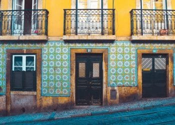 Una scena di strada colorata presenta un edificio giallo con intricati balconi in ferro battuto e pareti di piastrelle a motivi geometrici in blu e verde. L'edificio comprende una porta in legno scuro, una porta in vetro e finestre con persiane. La strada di fronte è acciottolata.