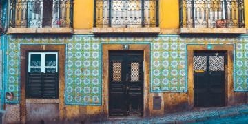 Una scena di strada colorata presenta un edificio giallo con intricati balconi in ferro battuto e pareti di piastrelle a motivi geometrici in blu e verde. L'edificio comprende una porta in legno scuro, una porta in vetro e finestre con persiane. La strada di fronte è acciottolata.