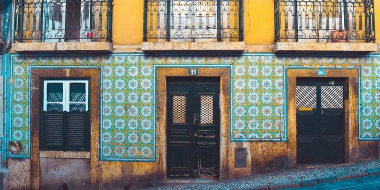 Una scena di strada colorata presenta un edificio giallo con intricati balconi in ferro battuto e pareti di piastrelle a motivi geometrici in blu e verde. L'edificio comprende una porta in legno scuro, una porta in vetro e finestre con persiane. La strada di fronte è acciottolata.