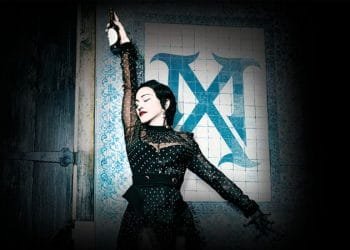 Una donna con i capelli scuri, che indossa un abito nero con paillettes, posa in modo drammatico con un braccio alzato mentre tiene in mano una bottiglia. È in piedi contro un muro piastrellato con una grande decorazione a "X" blu. La scena è scarsamente illuminata con un riflettore che la mette in risalto.