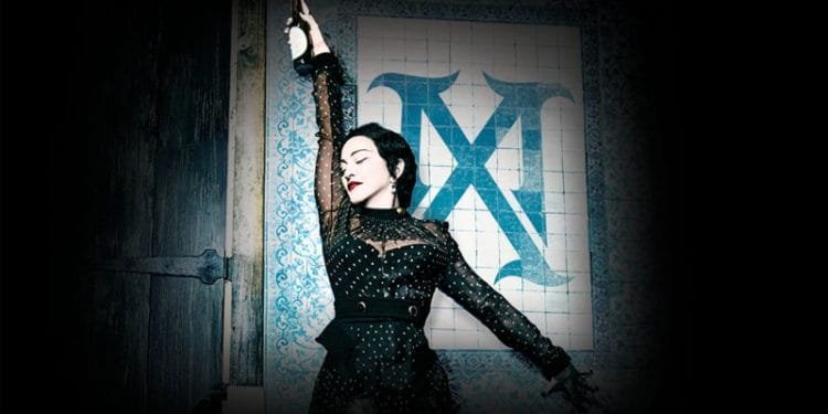 Una donna con i capelli scuri, che indossa un abito nero con paillettes, posa in modo drammatico con un braccio alzato mentre tiene in mano una bottiglia. È in piedi contro un muro piastrellato con una grande decorazione a "X" blu. La scena è scarsamente illuminata con un riflettore che la mette in risalto.