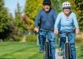 Una coppia di anziani va in bicicletta fianco a fianco su un sentiero asfaltato in un parco. Entrambi indossano caschi e maglioni e sorridono mentre si godono la giornata di sole. Lo sfondo presenta erba verde e alberi con fogliame autunnale.
