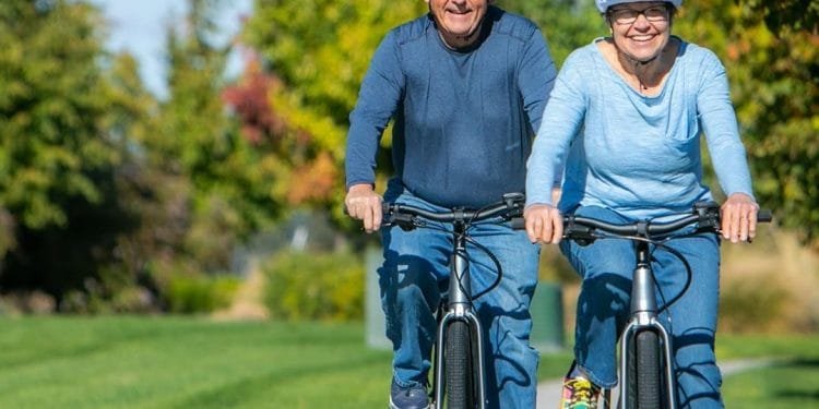 Una coppia di anziani va in bicicletta fianco a fianco su un sentiero asfaltato in un parco. Entrambi indossano caschi e maglioni e sorridono mentre si godono la giornata di sole. Lo sfondo presenta erba verde e alberi con fogliame autunnale.