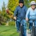 Una coppia di anziani va in bicicletta fianco a fianco su un sentiero asfaltato in un parco. Entrambi indossano caschi e maglioni e sorridono mentre si godono la giornata di sole. Lo sfondo presenta erba verde e alberi con fogliame autunnale.