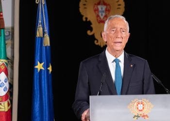 Un uomo anziano in giacca e cravatta sta su un podio mentre tiene un discorso. Dietro di lui ci sono le bandiere del Portogallo e dell'Unione Europea, insieme a un emblema ornato con lo stemma portoghese.