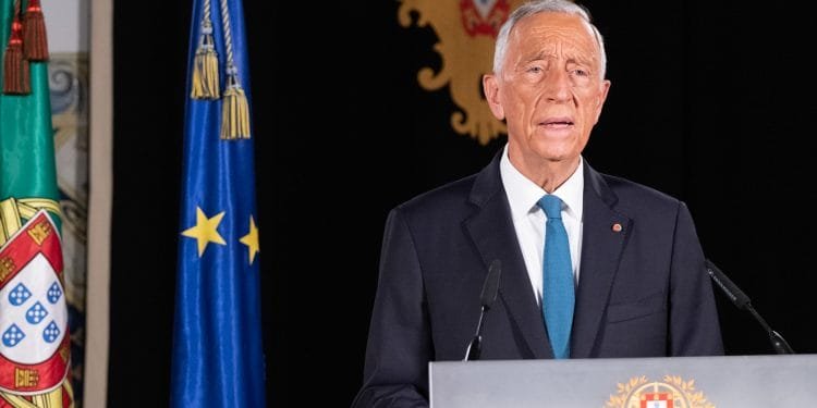 Un uomo anziano in giacca e cravatta sta su un podio mentre tiene un discorso. Dietro di lui ci sono le bandiere del Portogallo e dell'Unione Europea, insieme a un emblema ornato con lo stemma portoghese.