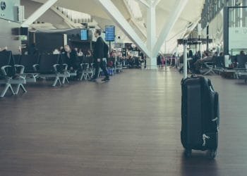 Una valigia nera si trova in primo piano in uno spazioso terminal aeroportuale. I passeggeri si siedono e camminano sullo sfondo vicino ai cancelli d'imbarco, con gli schermi delle partenze in alto e grandi travi strutturali visibili sul soffitto.