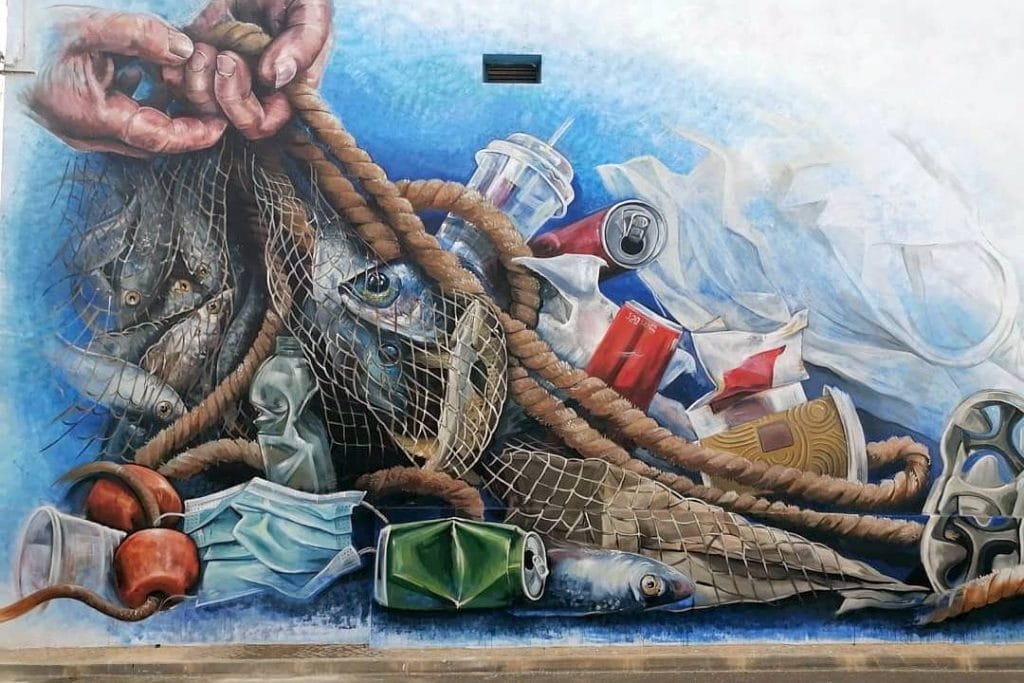 Un vivace murale di street art raffigura l’inquinamento marino, mostrando mani che tirano una rete da pesca piena di pesci e un inquietante mix di rifiuti di plastica come borse, bottiglie, tazze, lattine e maschere per il viso su uno sfondo blu. L’immagine evidenzia in modo toccante l’impatto ambientale delle attività umane sugli oceani.