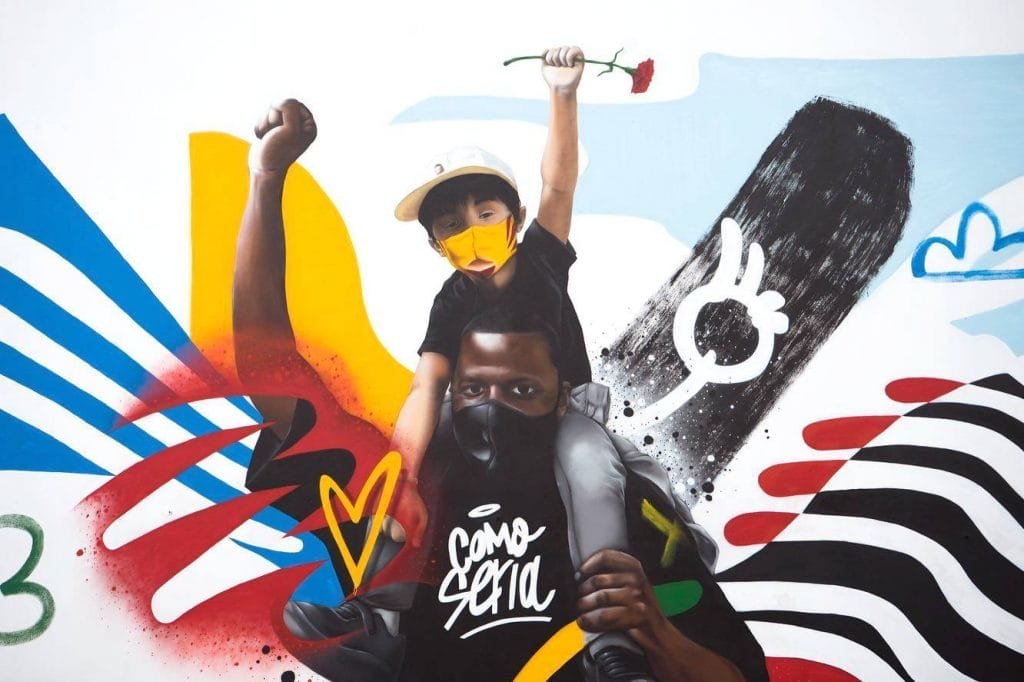 Un vivace murale di street art raffigura un bambino che indossa un berretto e una maschera gialla, che cavalca sulle spalle di un uomo con una maschera nera. Il bambino tiene in mano una rosa rossa. Forme e linee astratte colorate li circondano, con la frase "Cómo Será" scritta in stile graffiti bianco sotto.