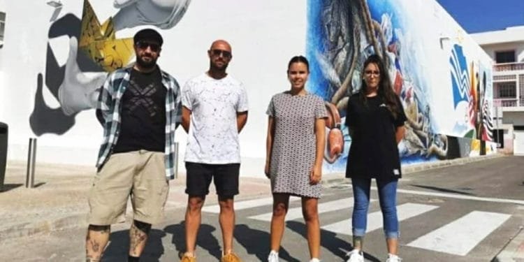 Quattro persone stanno con sicurezza su una strada di fronte a un grande murale caratterizzato da molteplici elementi artistici, tra cui una grande mano e disegni vivaci. Il gruppo è composto da tre uomini e una donna, tutti vestiti in modo casual. Il cielo è limpido e azzurro.