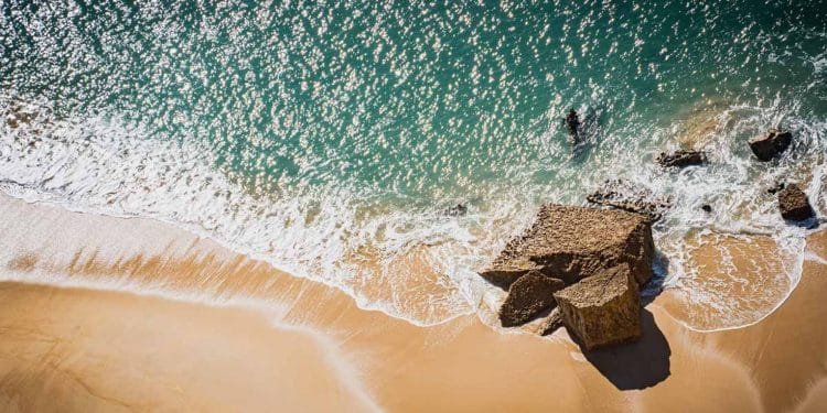 Veduta aerea di una spiaggia sabbiosa con onde turchesi che si infrangono dolcemente contro la riva. Grandi rocce sono parzialmente sommerse dall'acqua, creando una texture contrastante tra la sabbia liscia e le superfici ruvide delle rocce.
