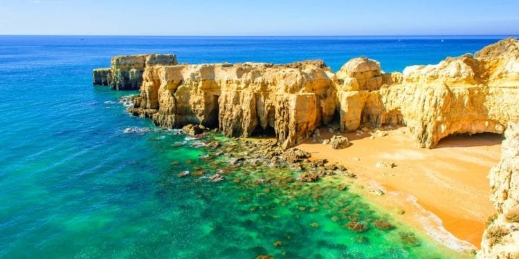 Una pittoresca scena costiera con spiagge di sabbia dorata, vivaci acque turchesi e spettacolari formazioni rocciose illuminate dal sole che si protendono nel mare sotto un cielo azzurro e limpido. Piccole onde lambiscono dolcemente la riva vicino ad un prominente affioramento roccioso.