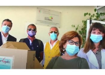 Un gruppo di cinque persone, quattro uomini e una donna, tutti con indosso le mascherine, stanno insieme in casa. Un uomo tiene in mano una grande scatola di cartone. Appaiono davanti a un cartello parzialmente visibile sullo sfondo, che suggerisce che si trovano in un contesto sanitario o organizzativo.