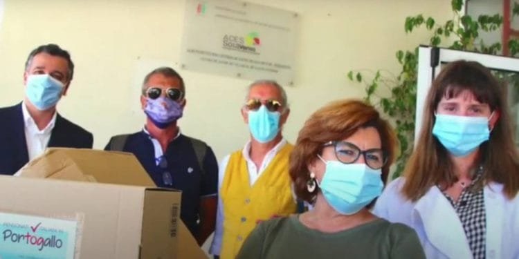 Un gruppo di cinque persone, quattro uomini e una donna, tutti con indosso le mascherine, stanno insieme in casa. Un uomo tiene in mano una grande scatola di cartone. Appaiono davanti a un cartello parzialmente visibile sullo sfondo, che suggerisce che si trovano in un contesto sanitario o organizzativo.