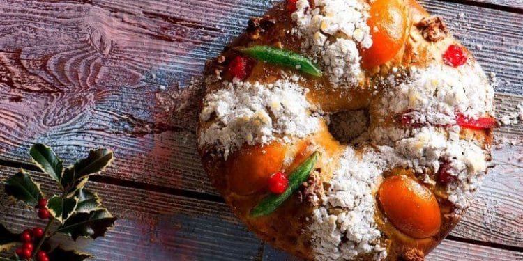 Un Bolo Rei festoso decorato con zucchero a velo, frutta candita e noci è posizionata su una superficie di legno rustica. Foglie e bacche di agrifoglio si trovano nell'angolo in basso a sinistra, esaltando il tema festivo di Natale.