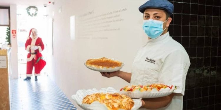 Una persona che indossa un berretto blu, un'uniforme bianca da chef e una maschera tiene in mano diversi dessert al piatto, tra cui una torta e alcuni pasticcini. Sullo sfondo, una persona vestita da Babbo Natale si trova vicino a una porta in una stanza con pavimento a scacchi bianchi e blu.