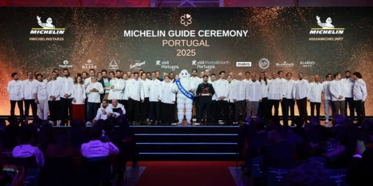Un folto gruppo di chef in giacca bianca è sul palco alla cerimonia delle premiazioni Guida Michelin Portogallo 2025. L' omino Michelin è al centro, circondato dai premiati. Lo sfondo mostra il logo Michelin e vari loghi degli sponsor, mentre il pubblico è seduto di fronte.