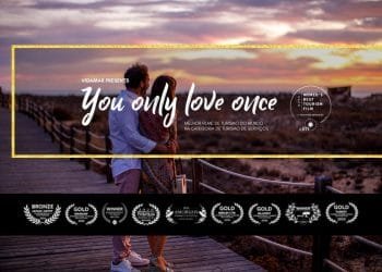 Una coppia si trova su un sentiero di legno che si affaccia su una spiaggia al tramonto. Il testo recita: "Vidamar presenta Ti ami solo una volta" e sottolinea che si tratta di un film turistico pluripremiato. Numerosi emblemi di premi sono allineati sul fondo.