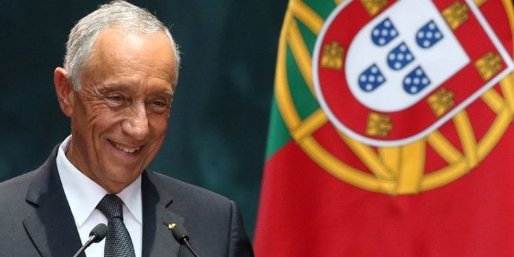 Un uomo sorridente in giacca e cravatta sta davanti ai microfoni, con una grande bandiera portoghese sullo sfondo.