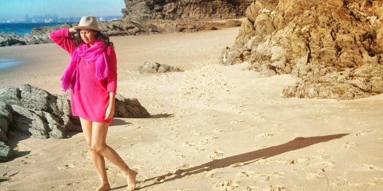 Una persona con un copricostume rosa brillante e un cappello beige si trova su una spiaggia sabbiosa circondata da formazioni rocciose. Sono scalzi, con una mano appoggiata sul cappello e proiettano una lunga ombra sulla sabbia. Il cielo è limpido e l'oceano è visibile sullo sfondo.