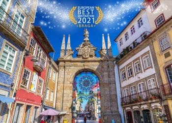 Una vivace strada a Braga, in Portogallo, è caratterizzata da edifici tradizionali colorati e un grande arco decorativo con dettagli decorati sotto un cielo azzurro brillante. Il testo "Migliore destinazione europea 2021, Braga" è visualizzato con scintillii sopra l'arco.