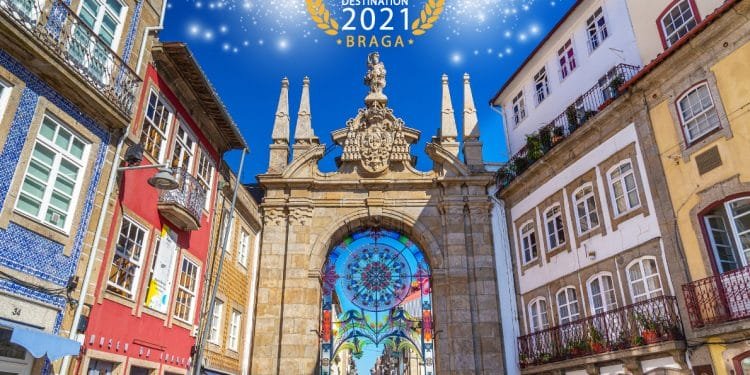 Una vivace strada a Braga, in Portogallo, è caratterizzata da edifici tradizionali colorati e un grande arco decorativo con dettagli decorati sotto un cielo azzurro brillante. Il testo "Migliore destinazione europea 2021, Braga" è visualizzato con scintillii sopra l'arco.