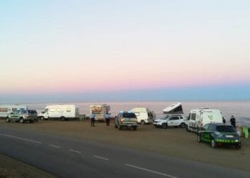 Camper e camper sono parcheggiati lungo una strada costiera al crepuscolo. Le persone stanno vicino alla costa e sono presenti diversi veicoli, tra cui un'auto della polizia. Il cielo passa dal rosa al blu man mano che la luce del giorno svanisce. Il mare calmo si estende fino all'orizzonte.