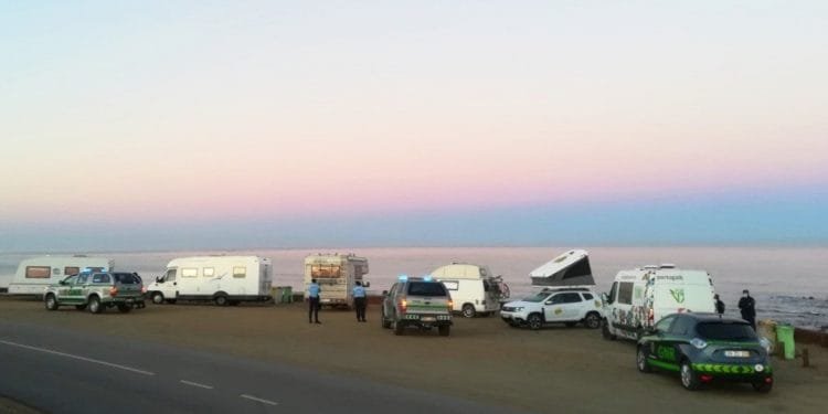 Camper e camper sono parcheggiati lungo una strada costiera al crepuscolo. Le persone stanno vicino alla costa e sono presenti diversi veicoli, tra cui un'auto della polizia. Il cielo passa dal rosa al blu man mano che la luce del giorno svanisce. Il mare calmo si estende fino all'orizzonte.